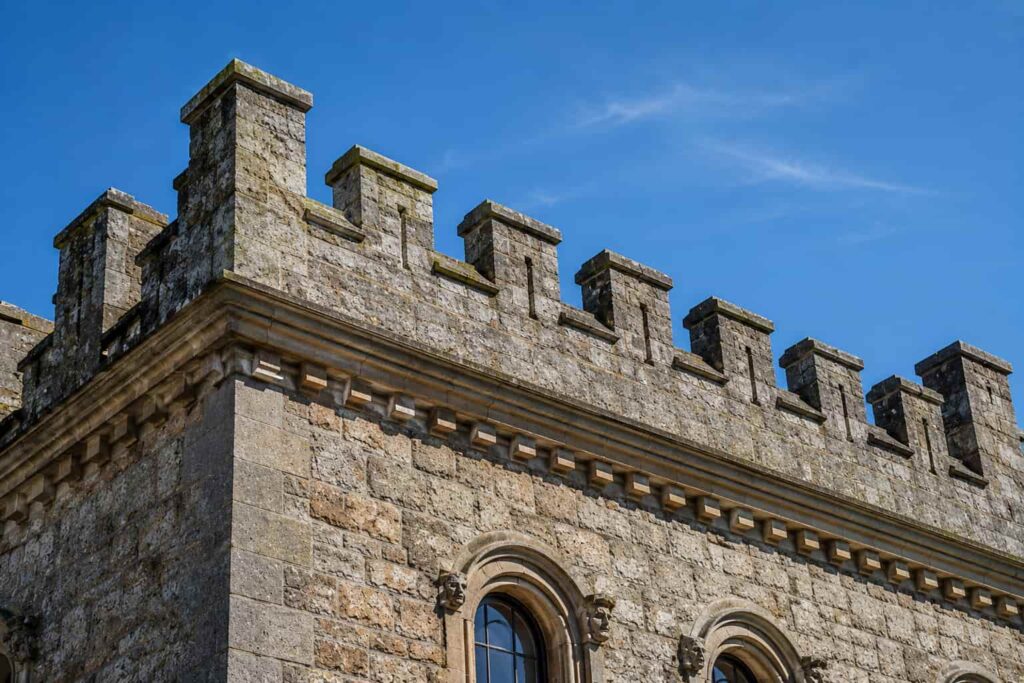 An embattled parapet wall featuring castle-style crenellations on a historic stone building with weathered masonry, decorative stonework, and a clear blue sky.