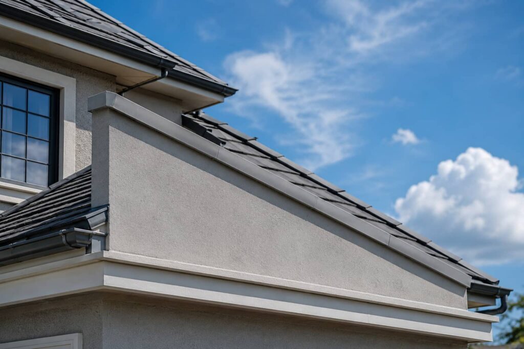 A close-up of a sloped concrete parapet wall on a modern residential building with a pitched roof, smooth plaster finish, concrete coping, roof shingles, gutters, and a clear blue sky