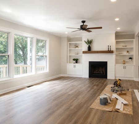 Bright modern living room during the final stage of interior renovation, featuring new wood flooring, white built-in shelves, a fireplace, large windows, and renovation tools neatly placed on the floor. interior renovation services