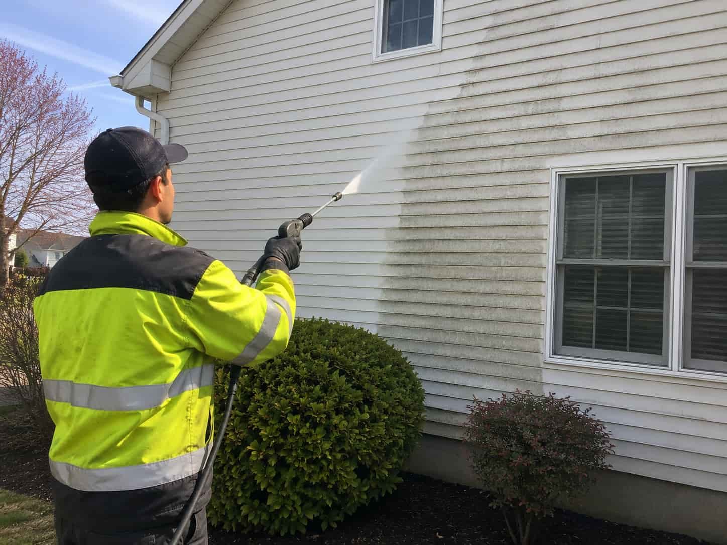 Professional worker wearing a safety jacket performing house pressure washing on a residential home in Maryland using commercial power washing equipment to remove dirt, mold, and mildew from exterior siding.